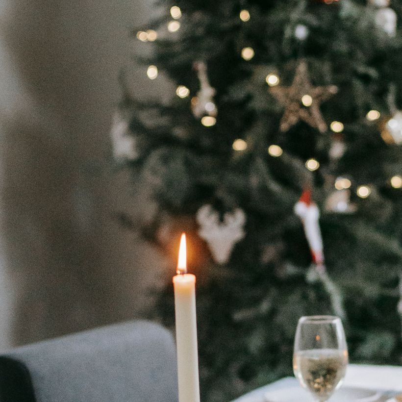 Table setting with burning candles and yummy dishes near fir tree with shiny garland during New Year holiday in house